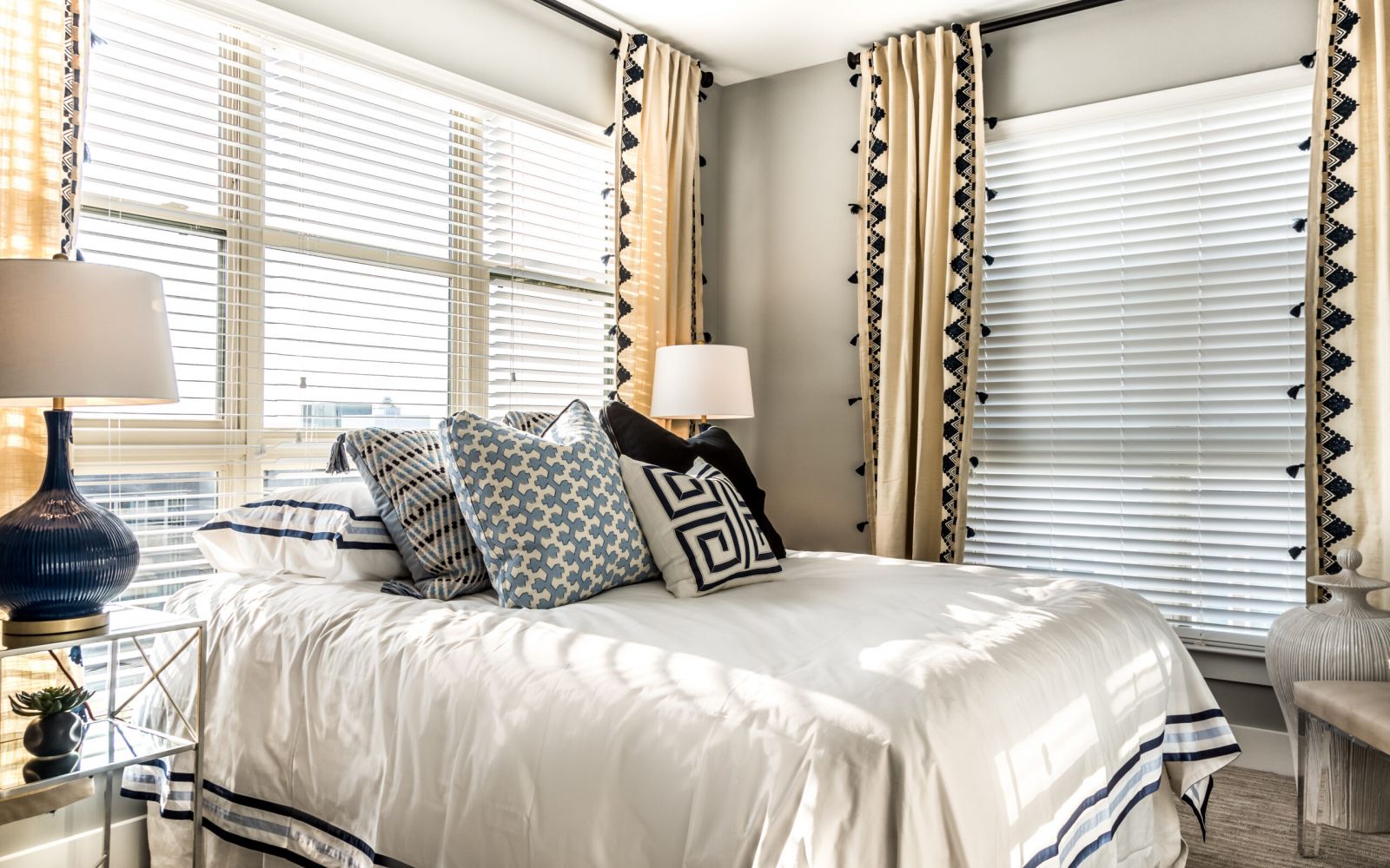 Bright bedroom with large windows, white bedding, patterned pillows, and beige curtains with black trim.