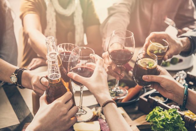 People clinking glasses and bottles in a toast around a table with food and drinks.
