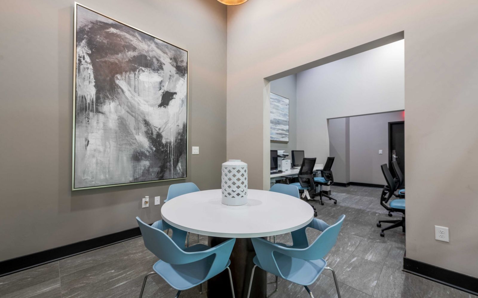 Round table with four blue chairs, large abstract painting, and office workspace in the background.
