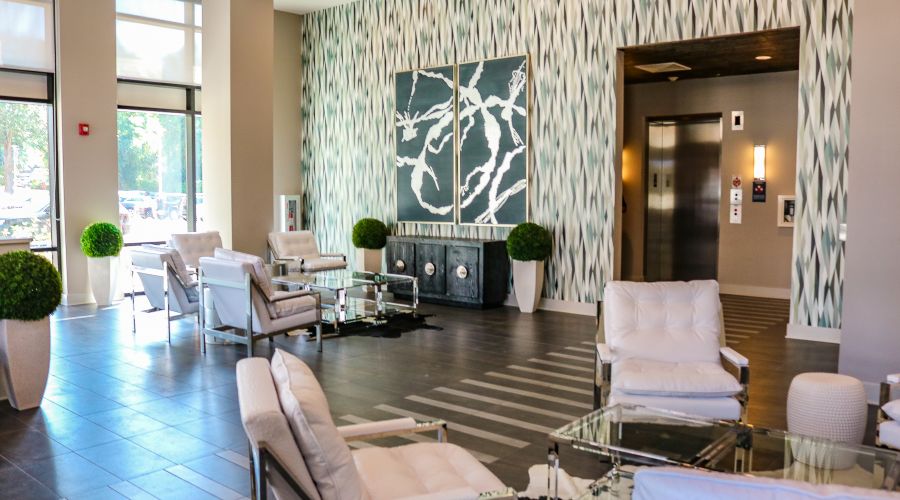 Modern lobby with white chairs, glass tables, elevator, decorative plants, and abstract wall art.
