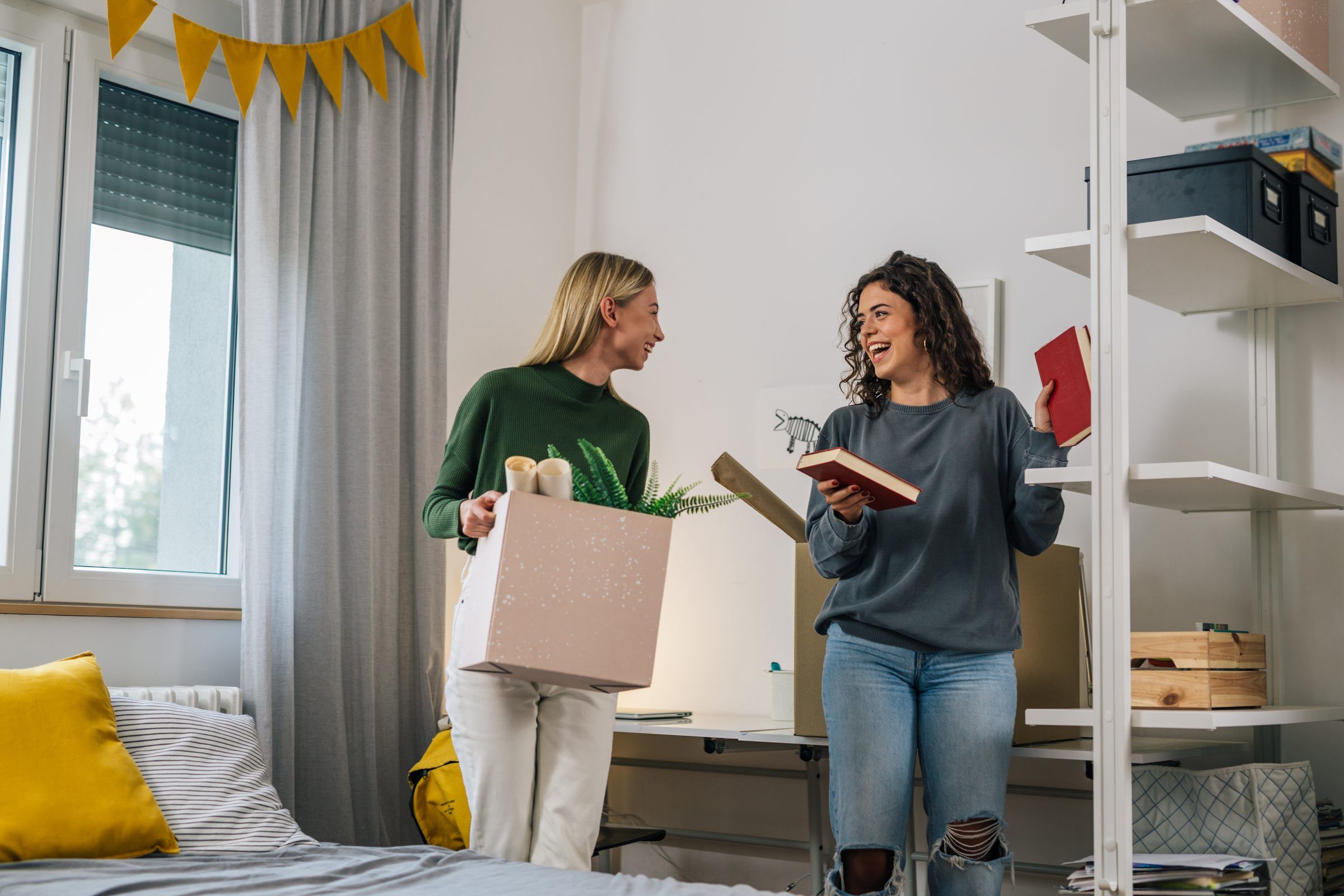 Two women smiling while unpacking boxes and organizing books in a bright bedroom or dorm room.