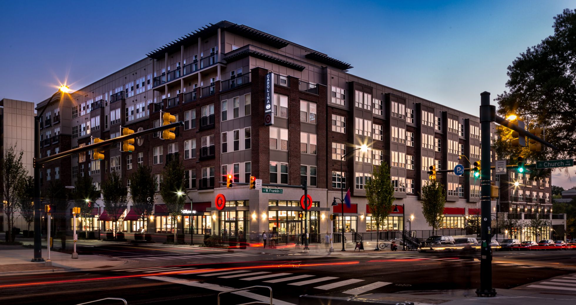 Modern apartment building with shops on the ground floor at dusk, streetlights and cars at the intersection.