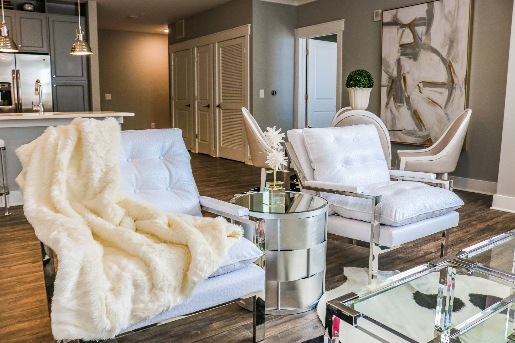 Modern living room with white chairs, a fur throw, round glass table, and abstract art on the wall.