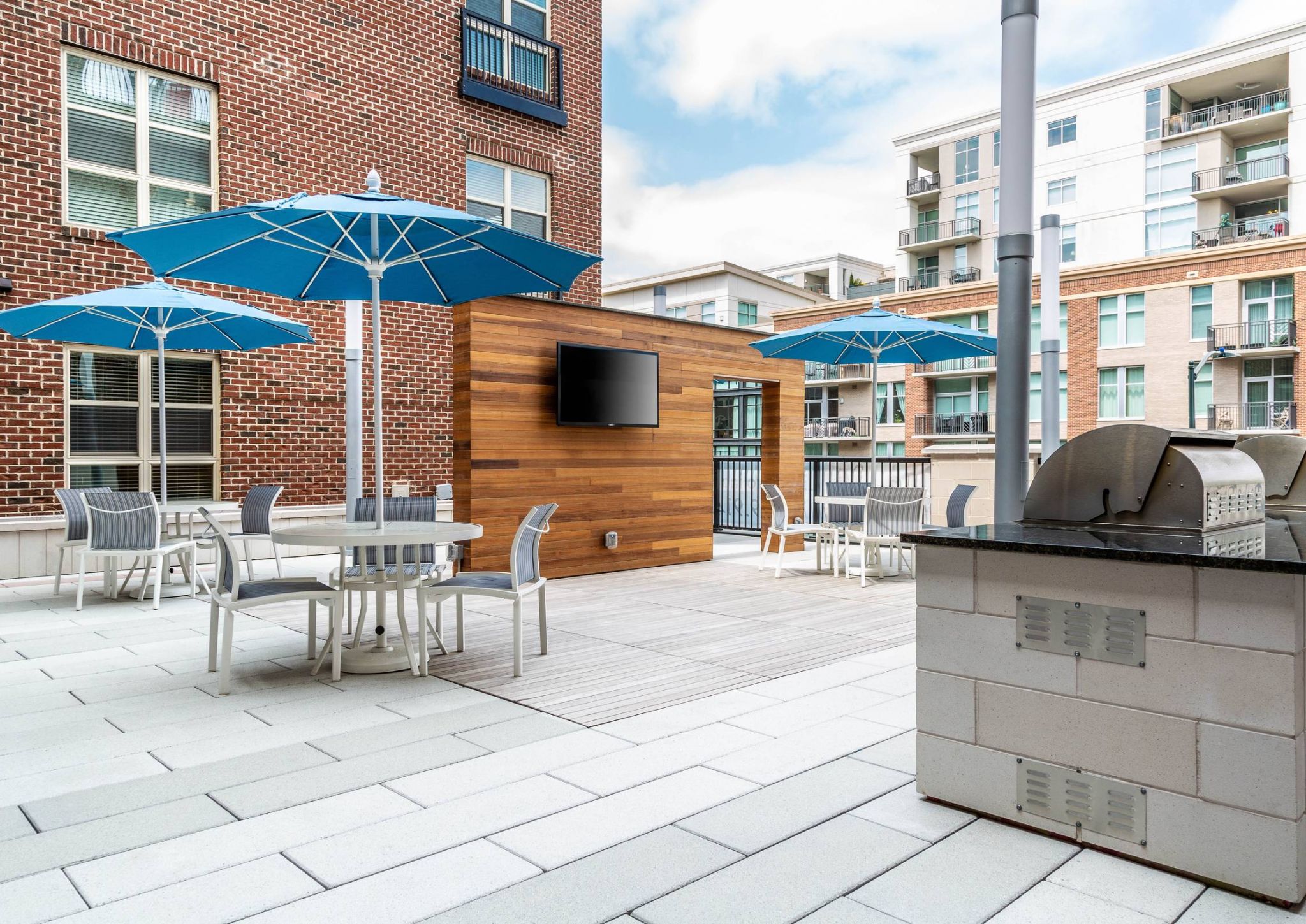 Modern outdoor patio with tables, blue umbrellas, a TV, and a grill, surrounded by apartment buildings.