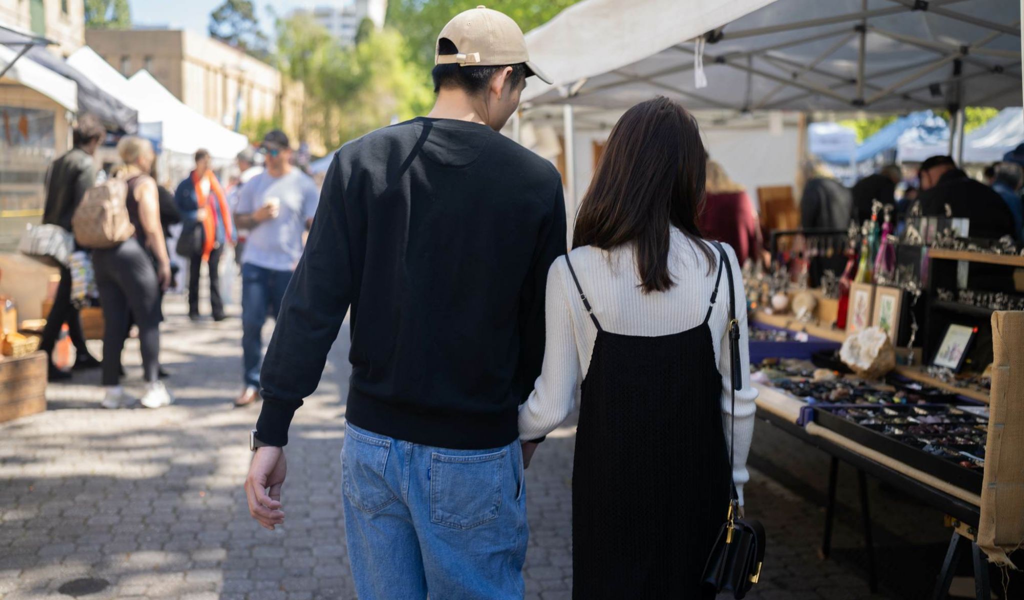 A couple walks arm-in-arm at an outdoor market with vendor stalls and shoppers around them.