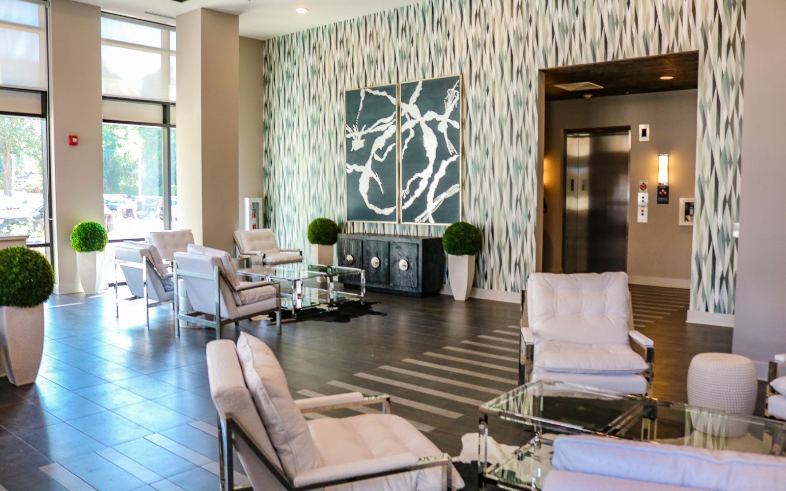 Modern lobby with white chairs, glass tables, elevator, decorative plants, and abstract wall art.