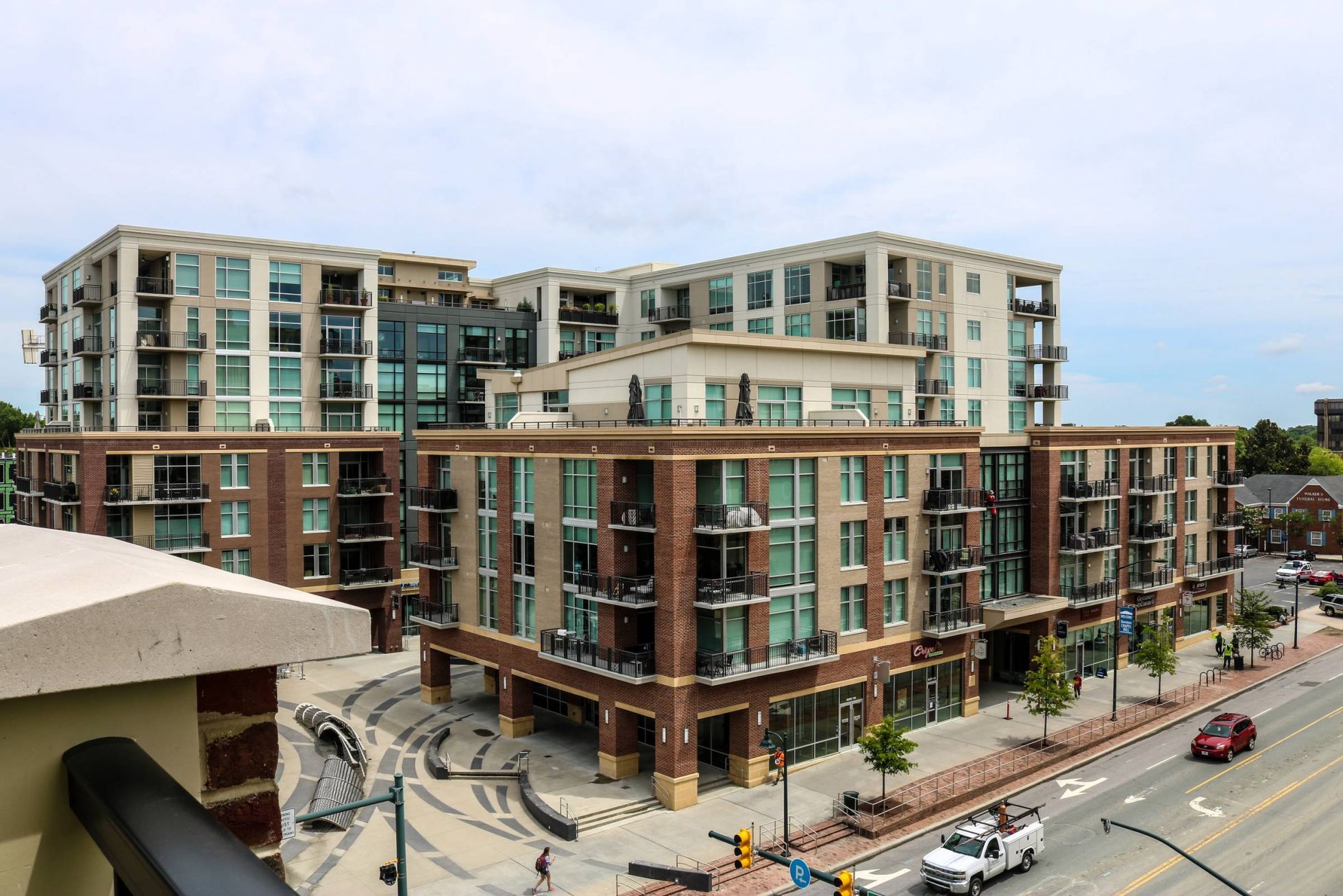 Modern mid-rise apartment complex with glass balconies, street-level shops, and a plaza alongside a city street.