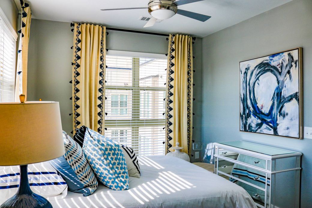 Bright bedroom with yellow curtains, blue abstract art, decorative pillows, and a ceiling fan at Carolina Square apartments