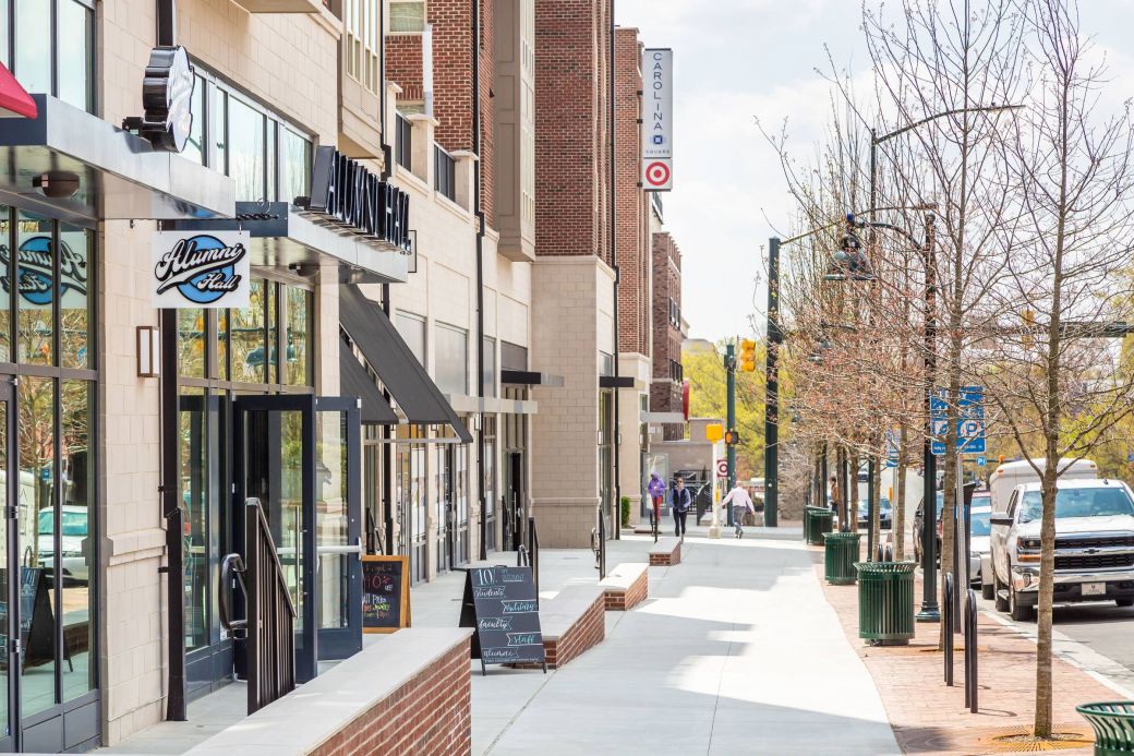 Modern city sidewalk with shops, a Target sign, parked cars, and a few pedestrians on a sunny day.