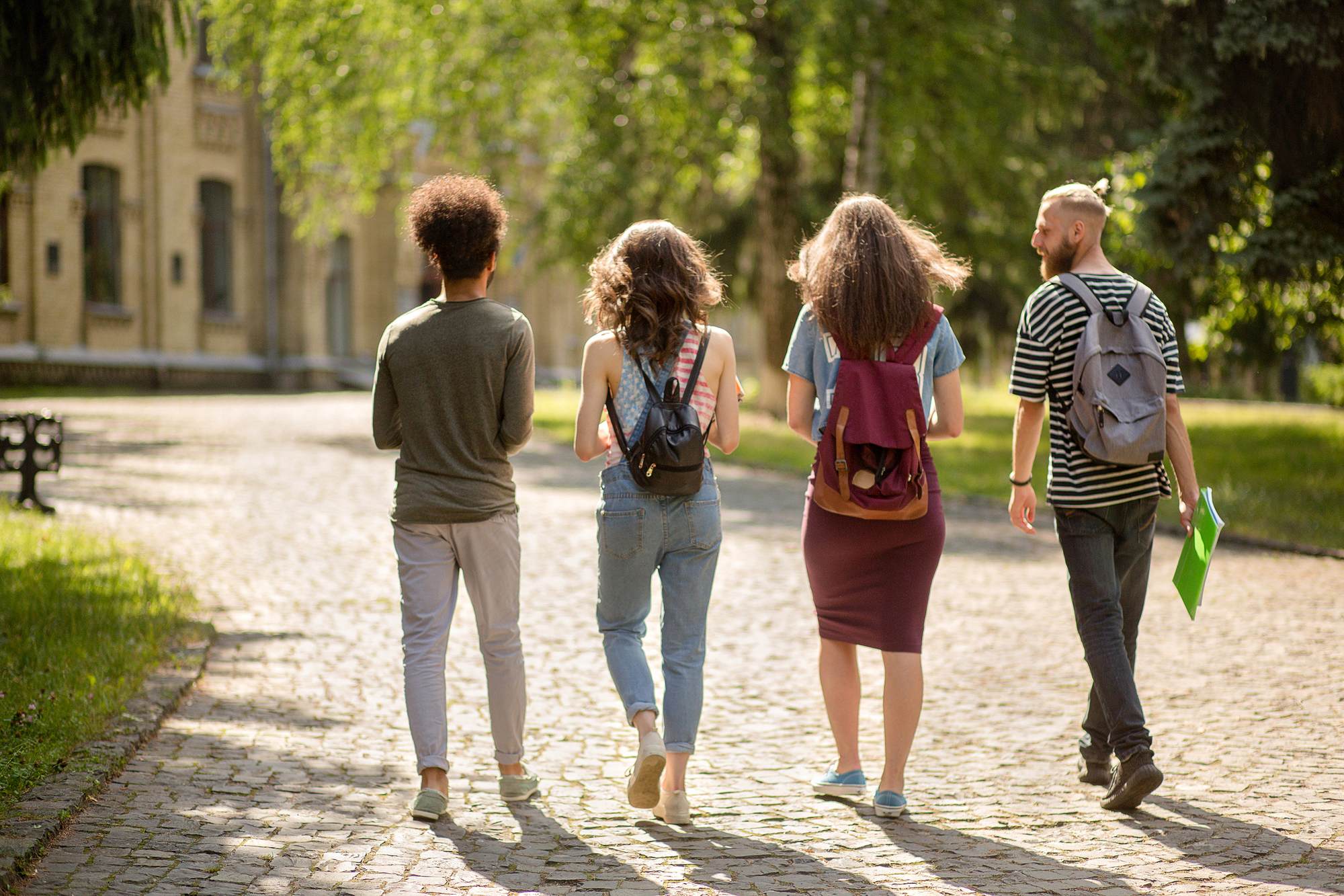 Four young adults with backpacks walk together on a sunny, tree-lined cobblestone path.