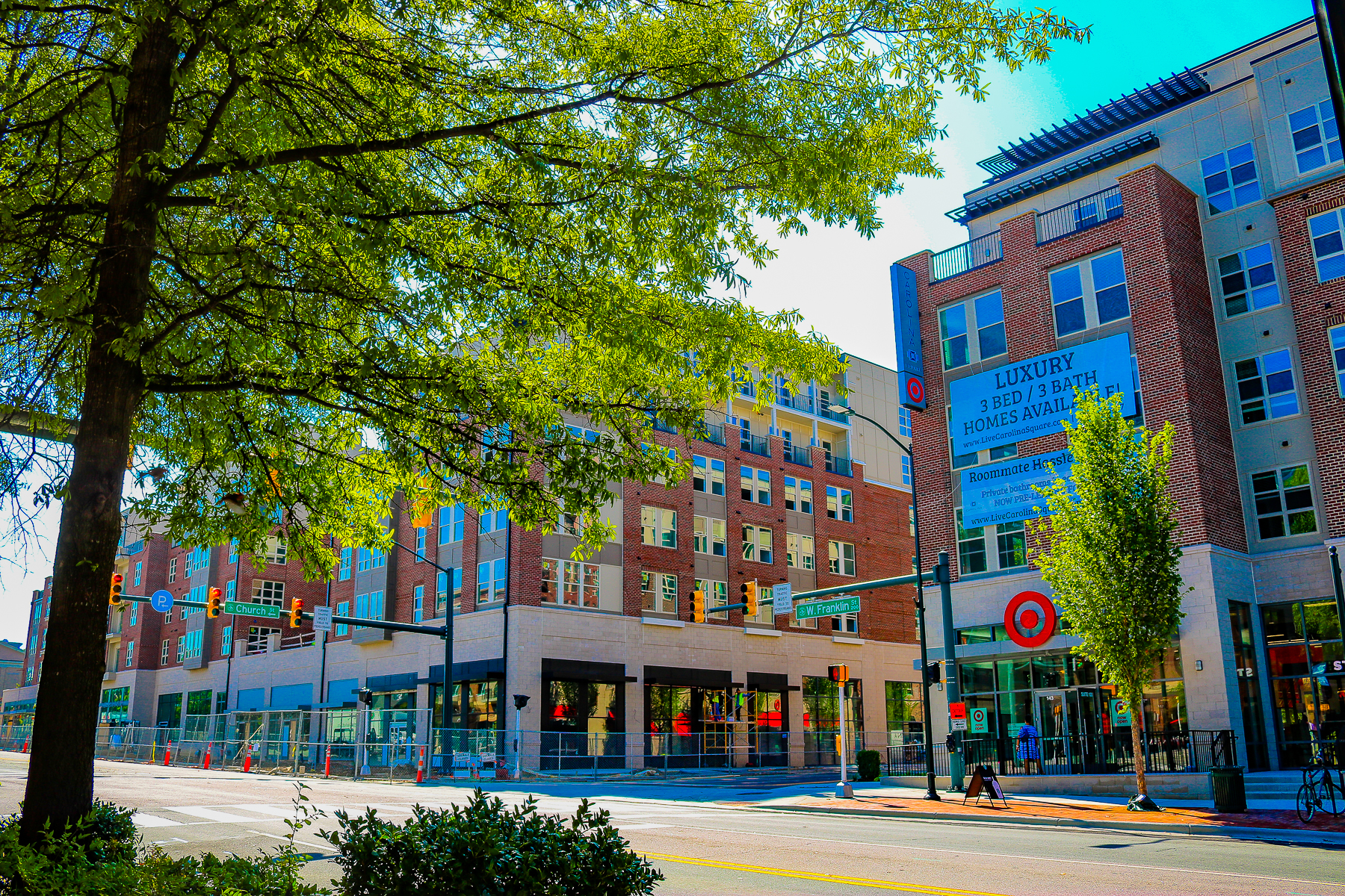 Sunny city street with modern apartment buildings, trees, and a sign advertising luxury homes for rent.
