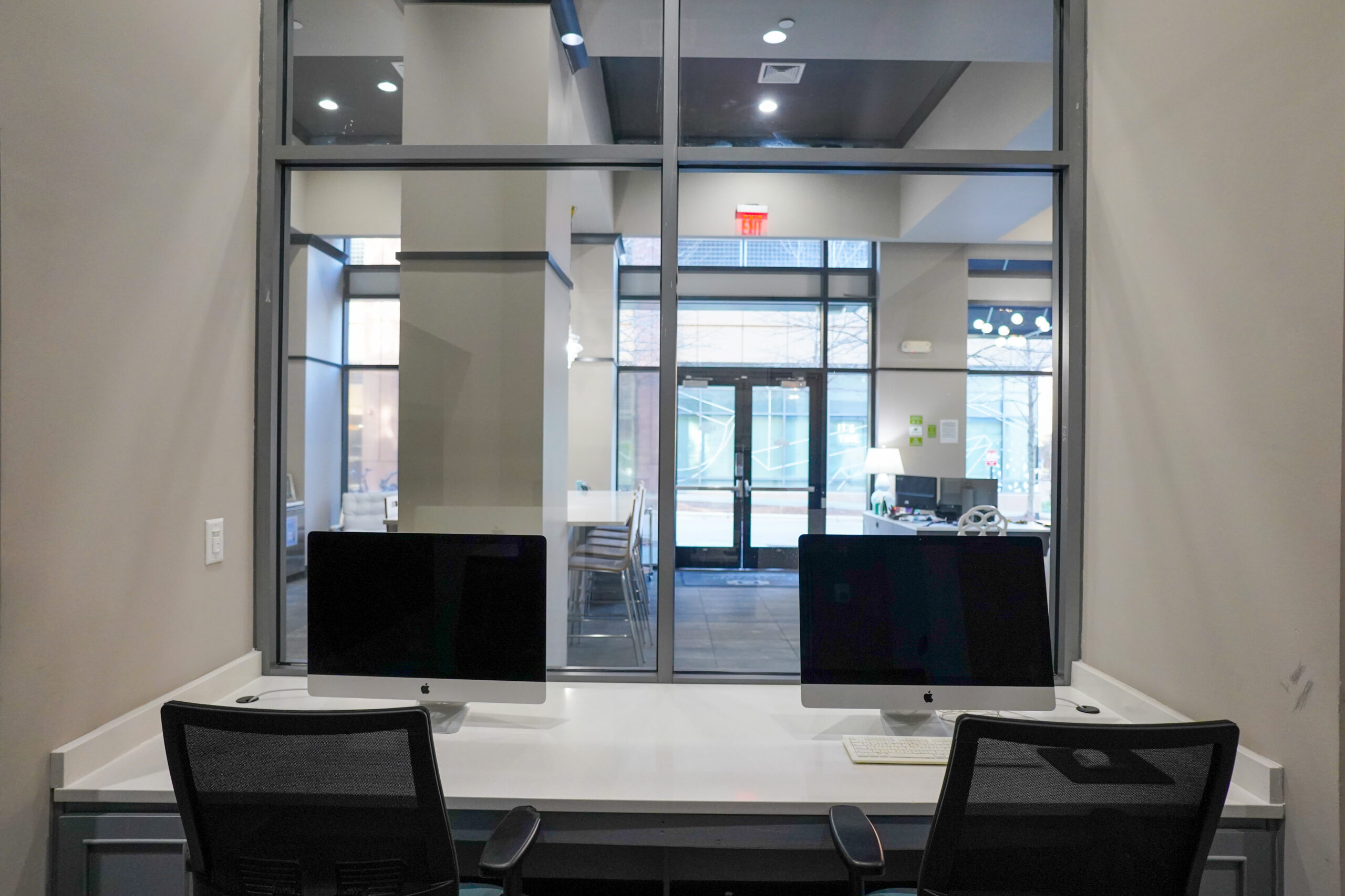 Two desktop computers on a desk in front of large windows inside a modern office space.