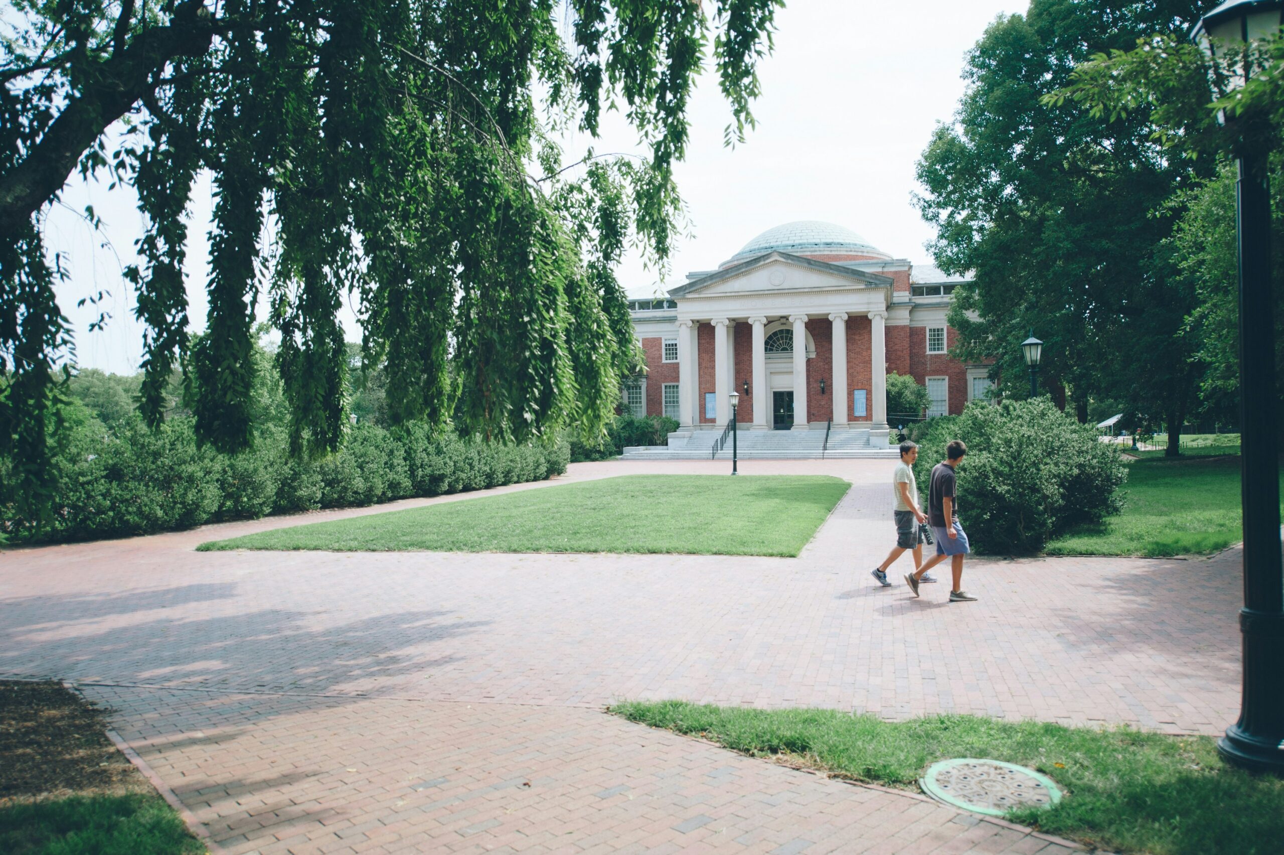 Two people walk on a brick path in front of a large, domed, brick building surrounded by trees.