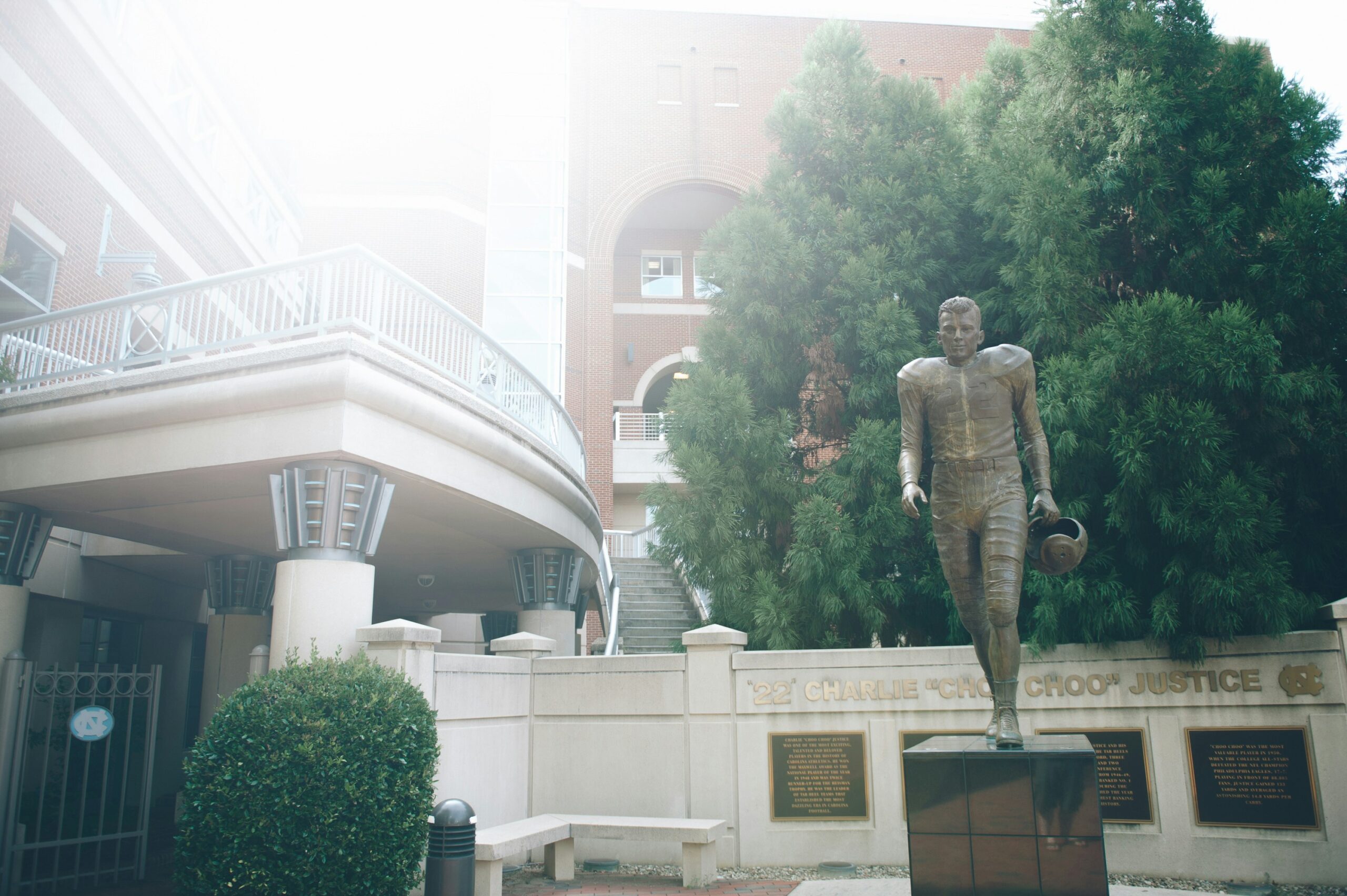 Bronze UNC football player statue holding helmet near a building and tall green tree on a sunny day.