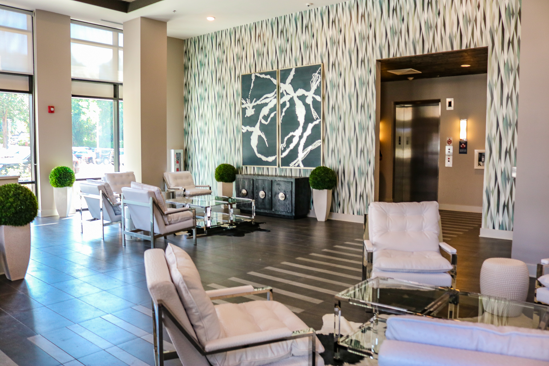 Modern lobby with white chairs, glass tables, elevator, decorative plants, and abstract wall art.