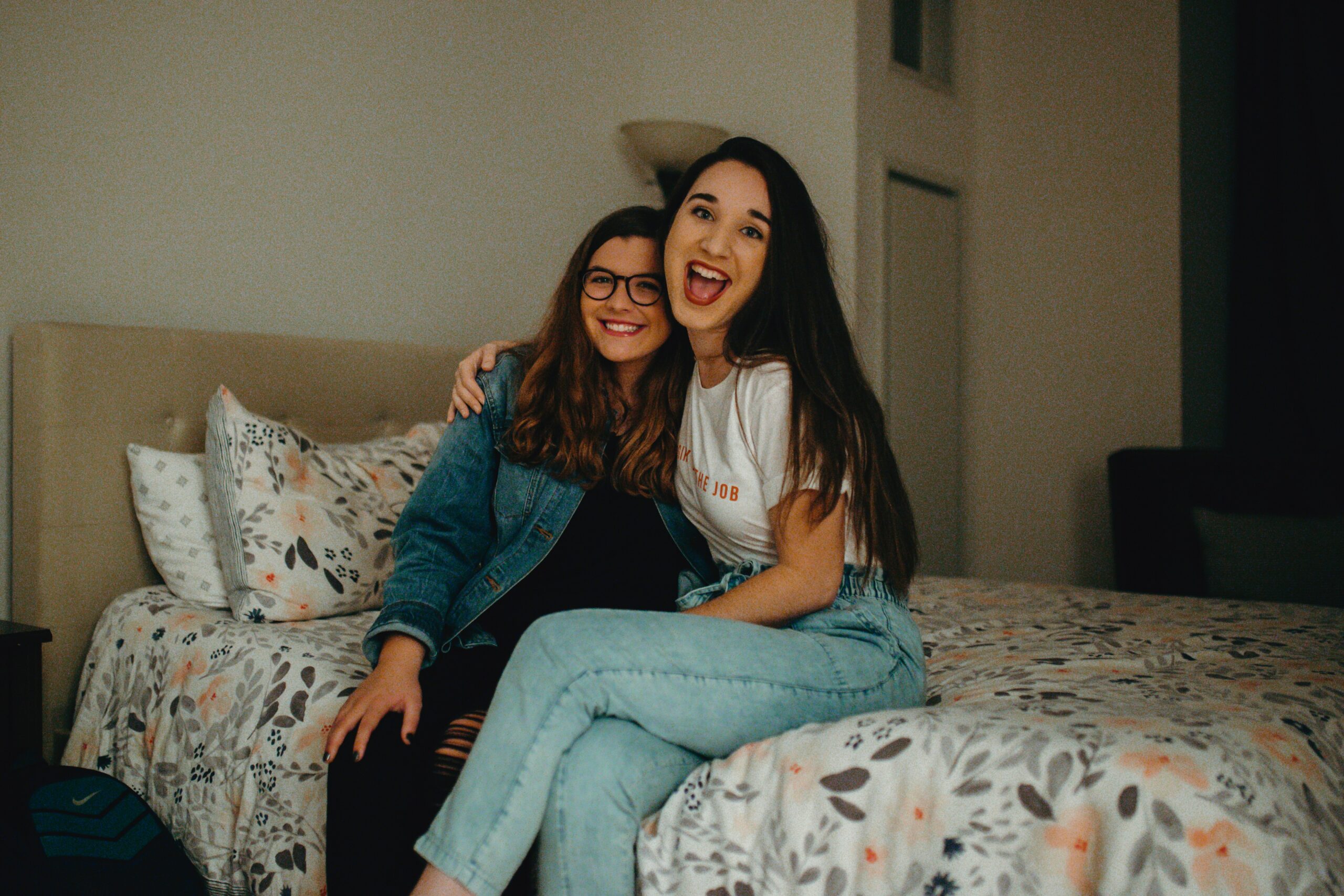 Two young women smiling and sitting together on a bed in a cozy, warmly lit Carolina Square bedroom—the perfect roommate vibe.