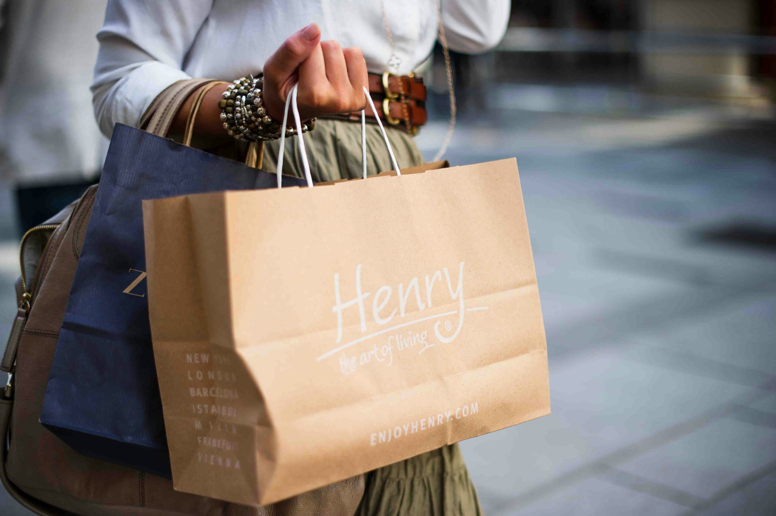 Person holding shopping bags, including a brown one labeled “Henry the art of living,” on a city street.