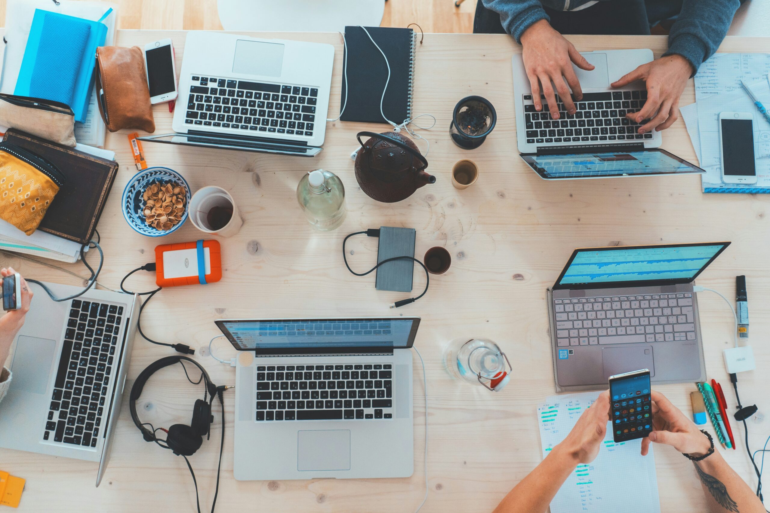 Overhead view of a cluttered work desk with laptops, notebooks, snacks, and people working together.