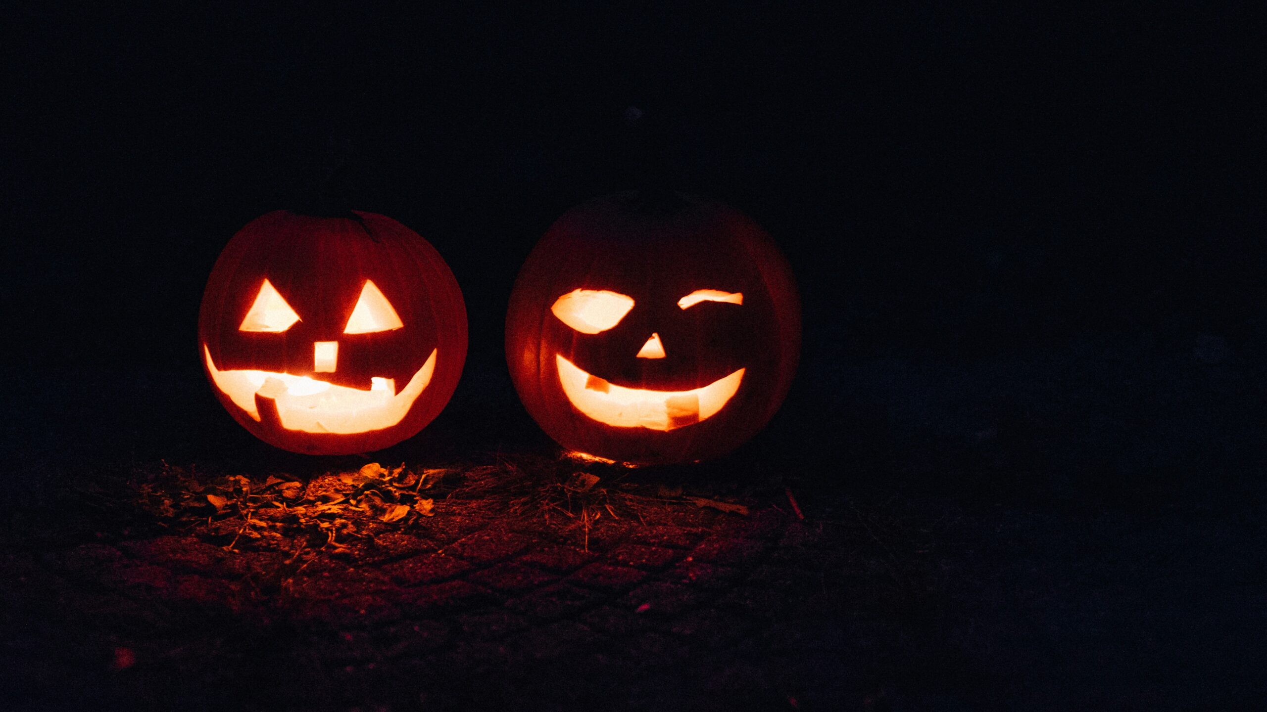 Two glowing jack-o’-lanterns with carved faces sit side by side on the ground in the dark.