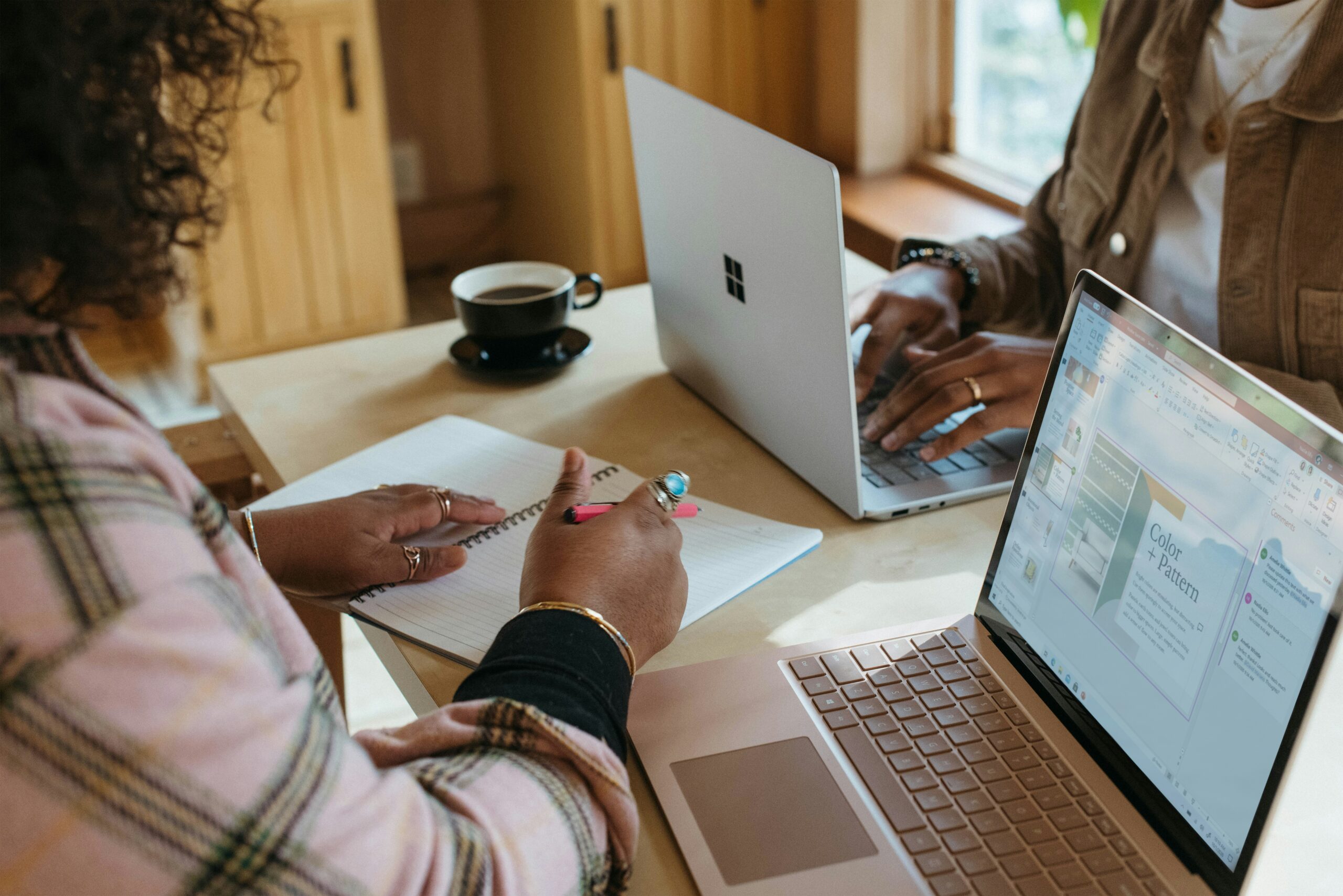 Two people working at a table with laptops and notebooks, one writing and the other typing.
