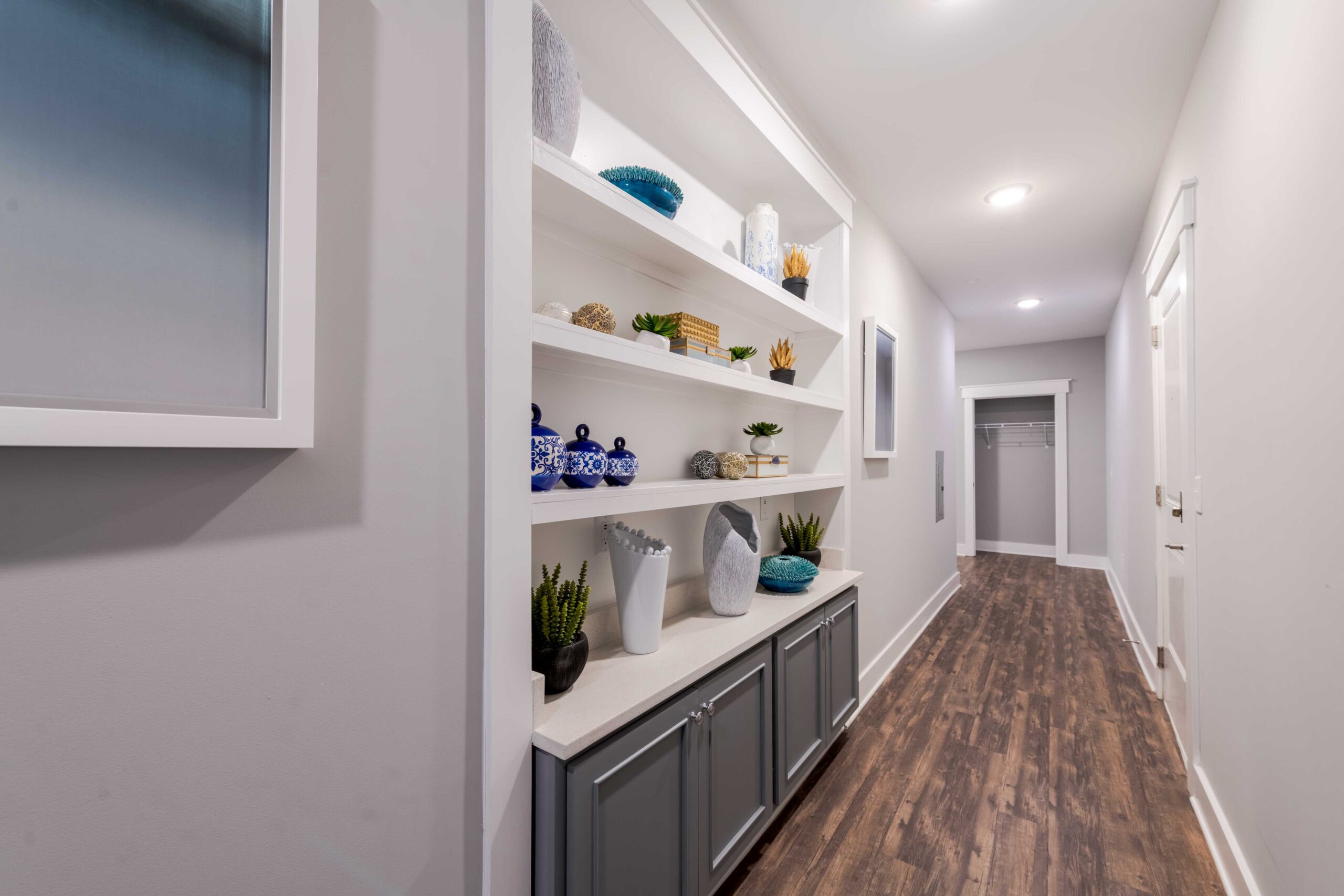 A hallway with wooden floors, built-in shelves holding decor, and an open closet at the end.