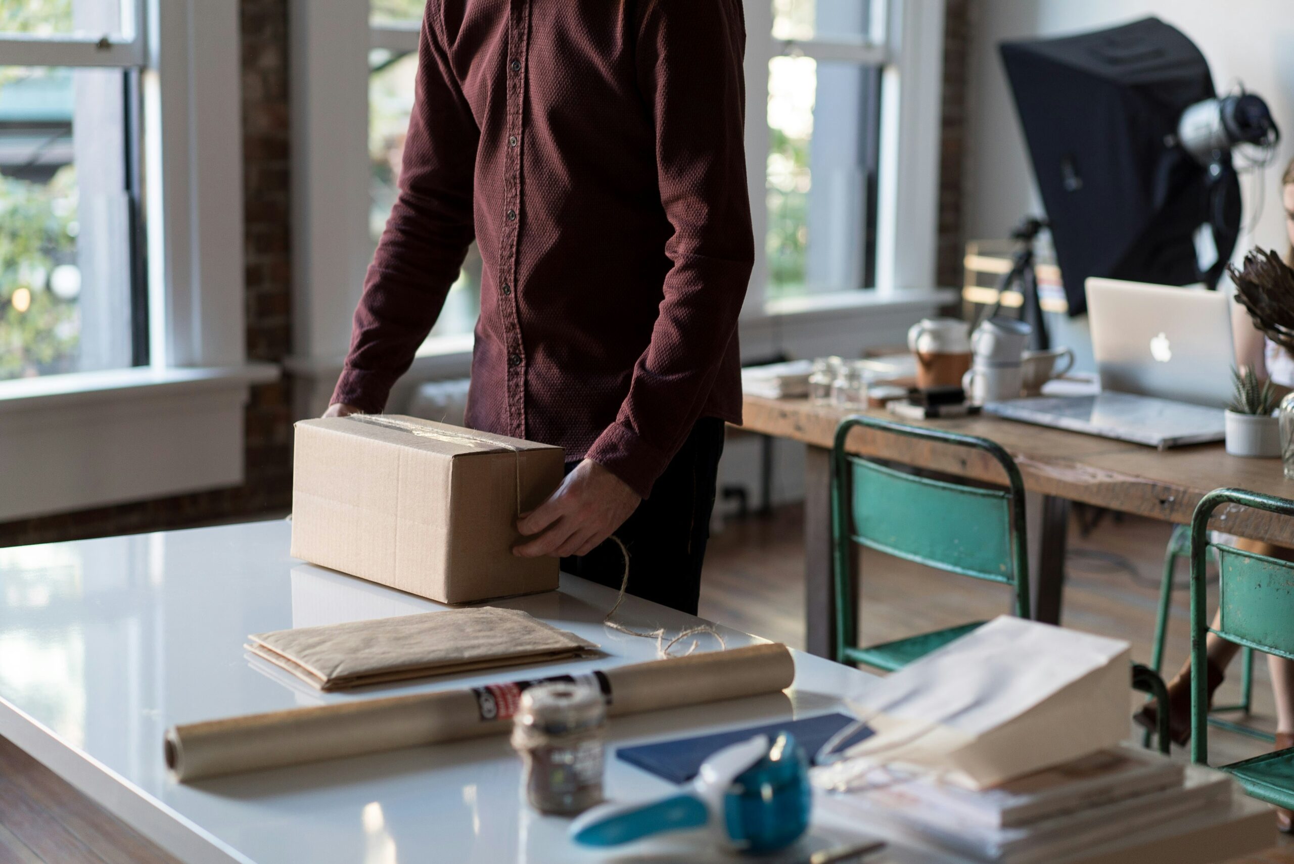Person in a maroon shirt packing a cardboard box on a white desk with office supplies nearby.