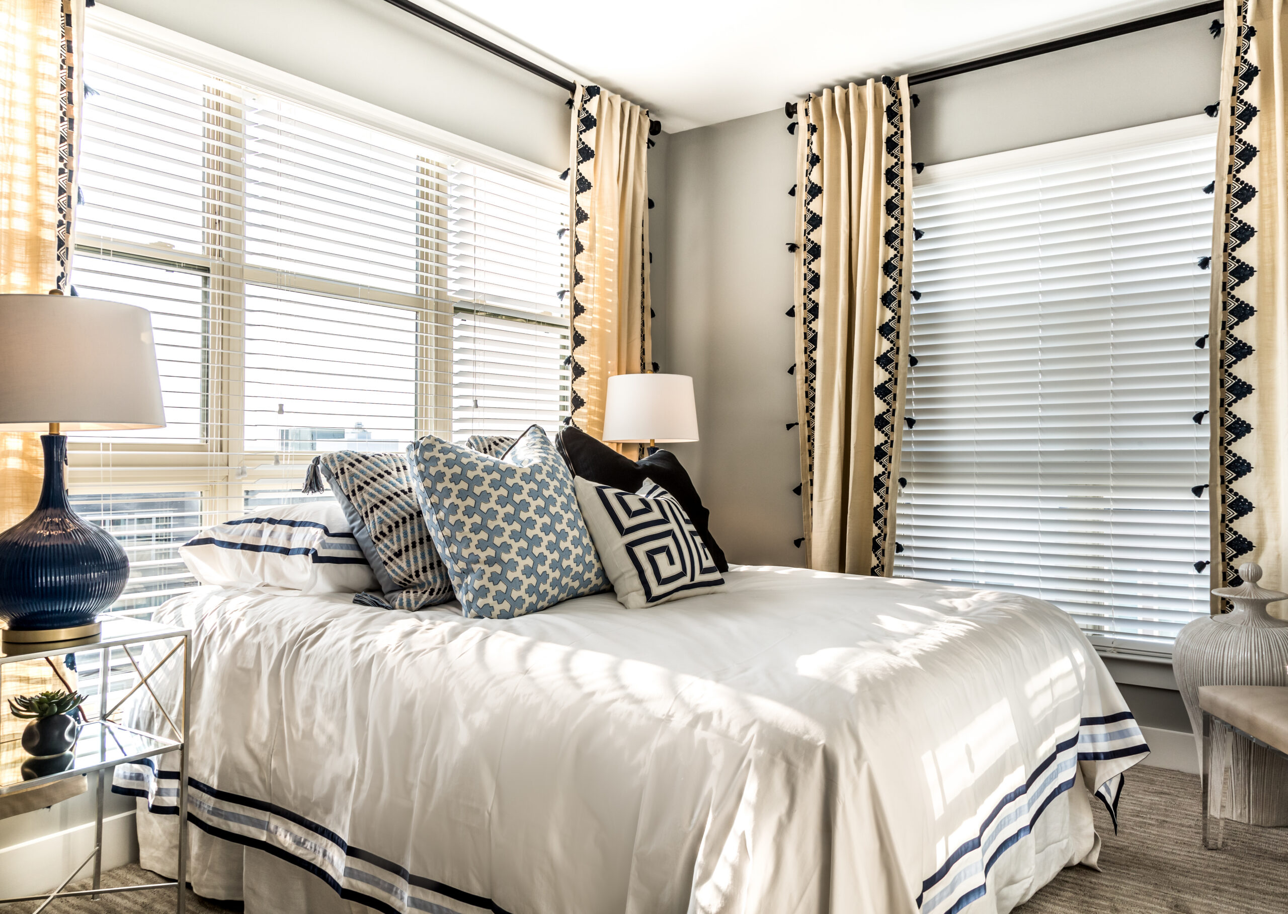 Bright bedroom with large windows, white bedding, patterned pillows, and beige curtains with black trim.