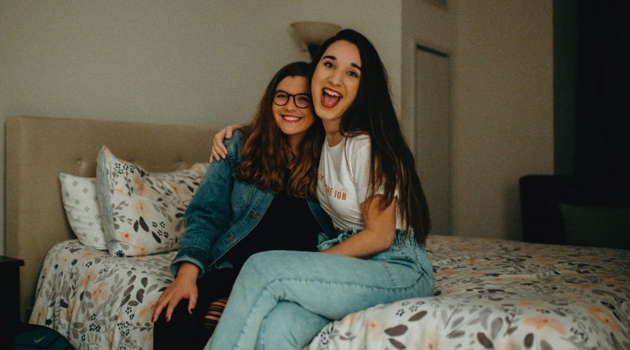 Two young women smiling and sitting together on a bed in a cozy, warmly lit Carolina Square bedroom—the perfect roommate vibe.