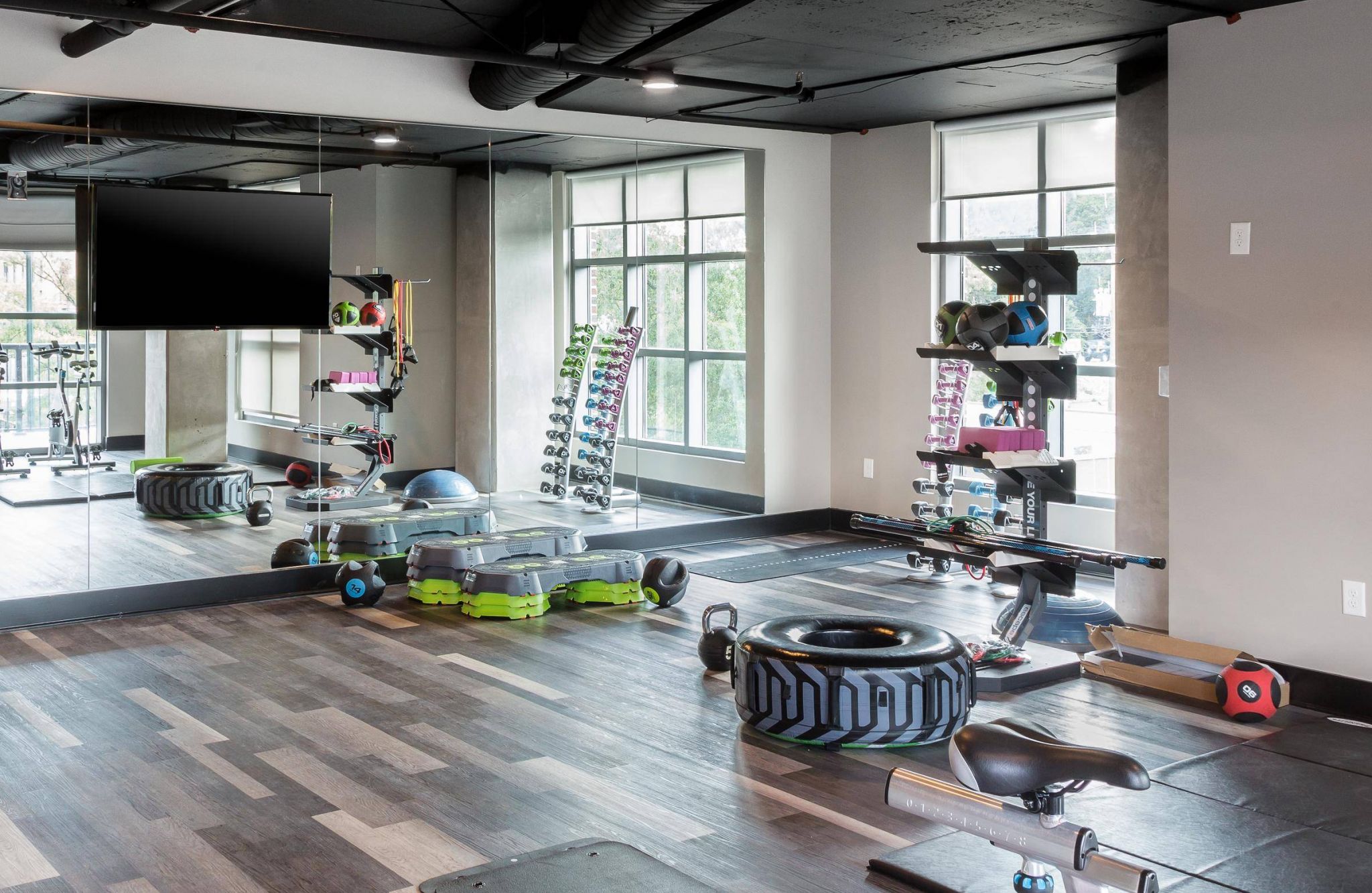 Modern gym with large mirrors, weights, exercise equipment, and natural light from big windows.
