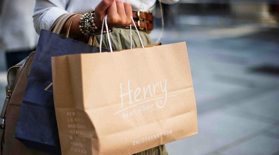 Person holding shopping bags, including a brown one labeled “Henry the art of living,” on a city street.