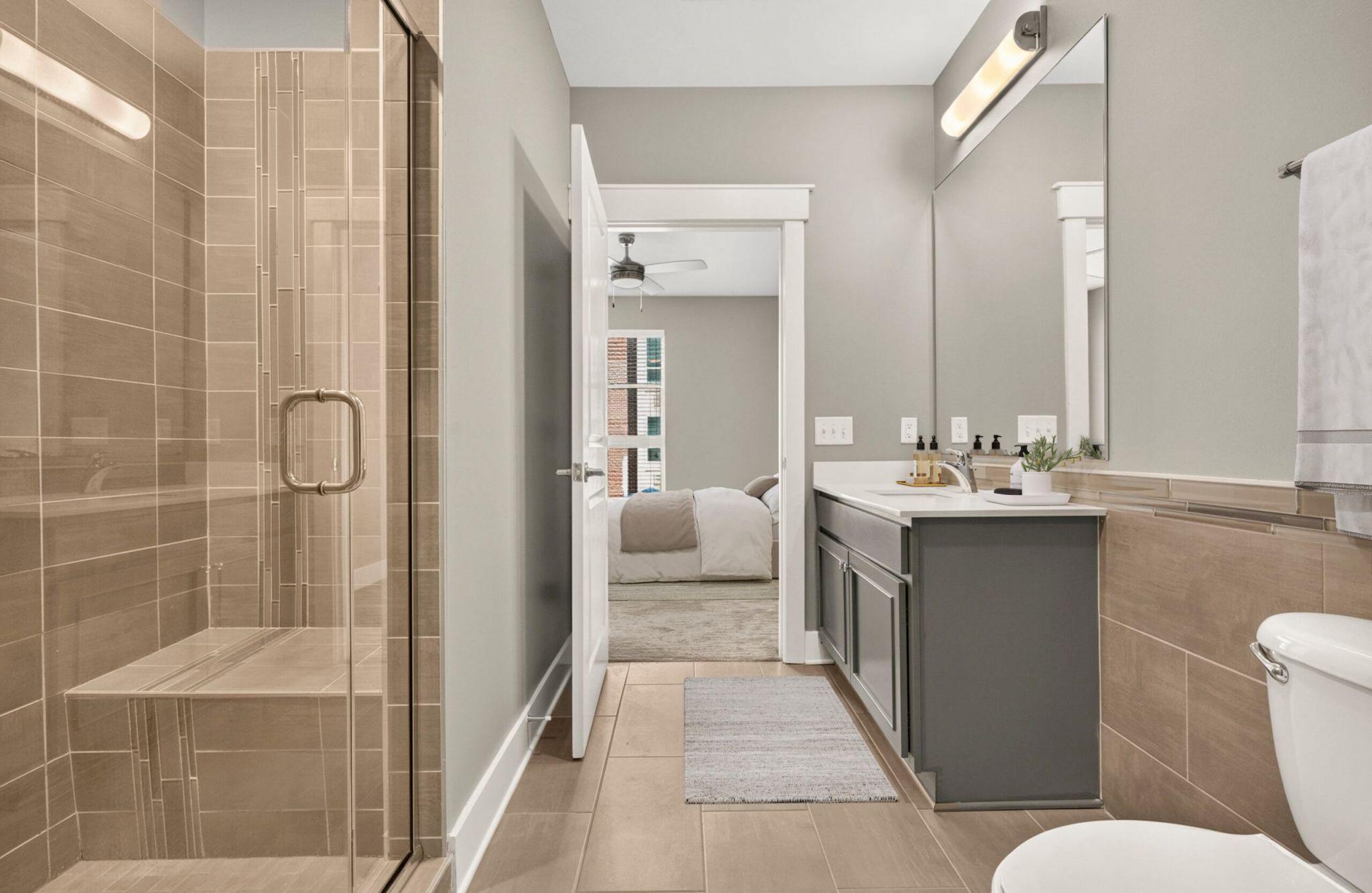 Modern bathroom with glass shower, large mirror, double sink, gray cabinets, and door open to a bedroom.