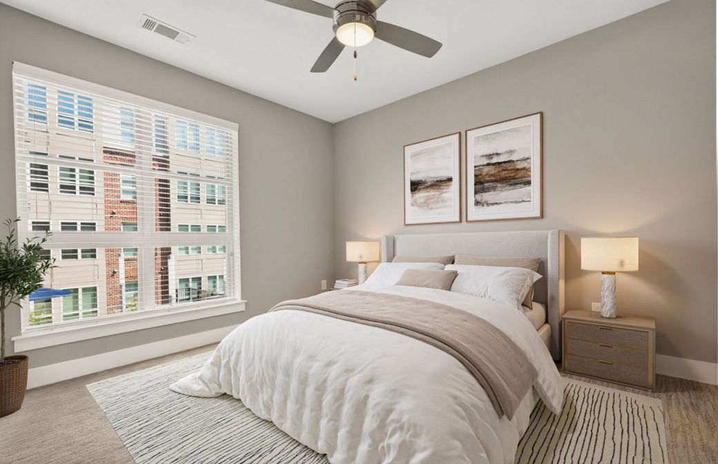 Modern bedroom with beige walls, large window, bed with white bedding, nightstands, lamps, art above headboard, and ceiling fan.