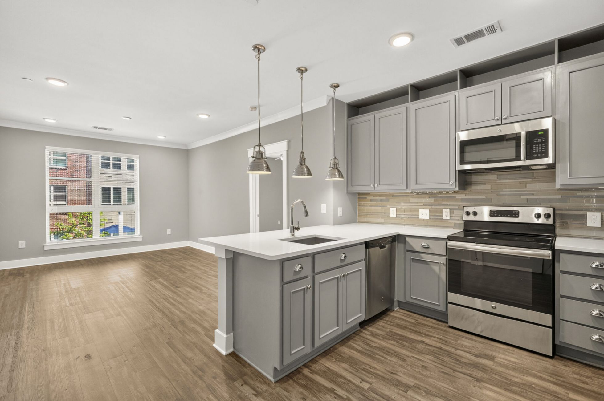 Modern kitchen with gray cabinets, stainless appliances, pendant lights, and open space with large window and wood flooring.