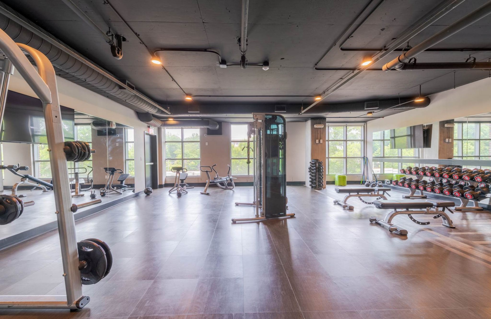 Spacious gym with weight racks, benches, exercise bikes, and large windows letting in natural light.
