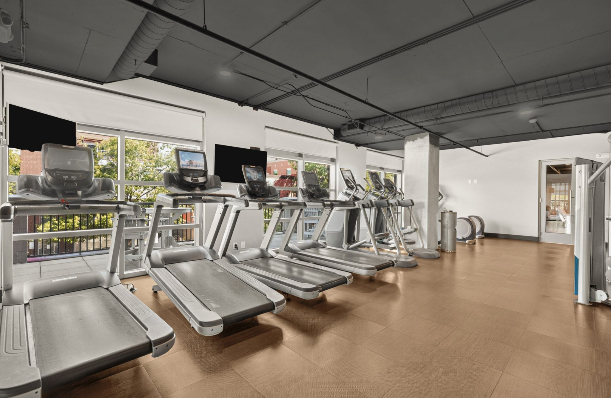 Row of treadmills facing large windows in a modern gym with brown floors and black ceiling.
