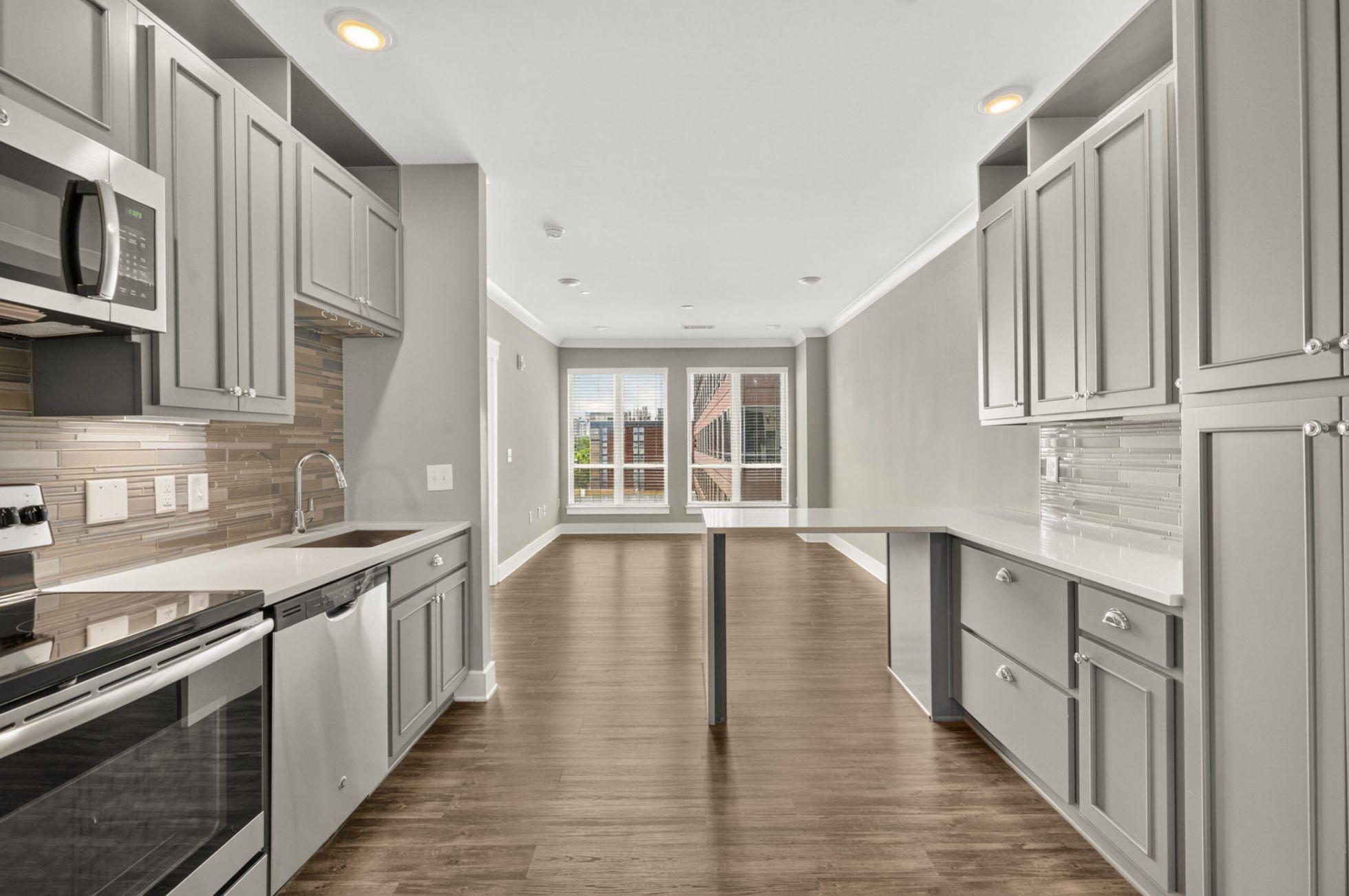 Modern kitchen with gray cabinets, white countertops, wood floor, and open view into a bright living area with large windows.