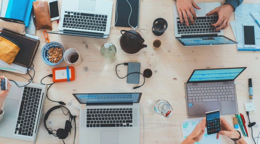 Overhead view of a cluttered work desk with laptops, notebooks, snacks, and people working together.