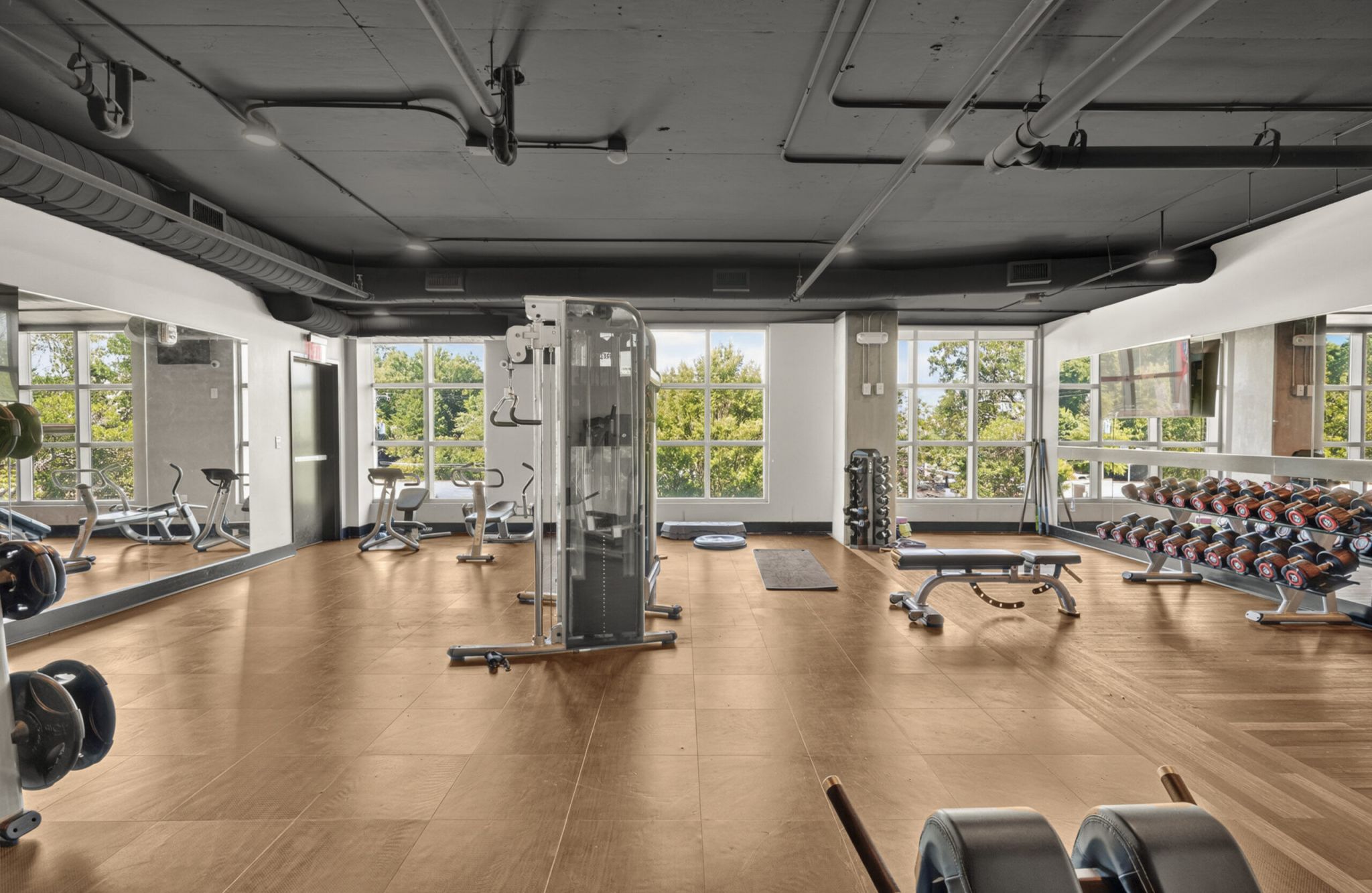 Spacious gym with large windows, weight racks, exercise machines, benches, and cardio equipment on a wood floor.