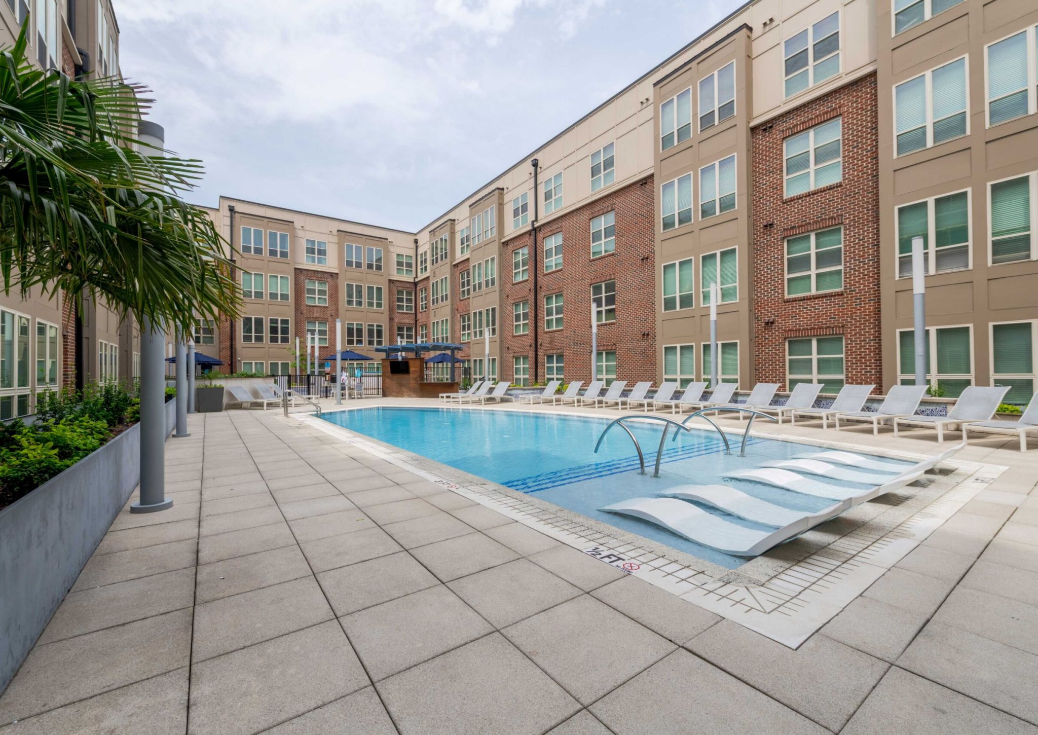 Modern apartment courtyard with a swimming pool, lounge chairs, and palm trees on a cloudy day.