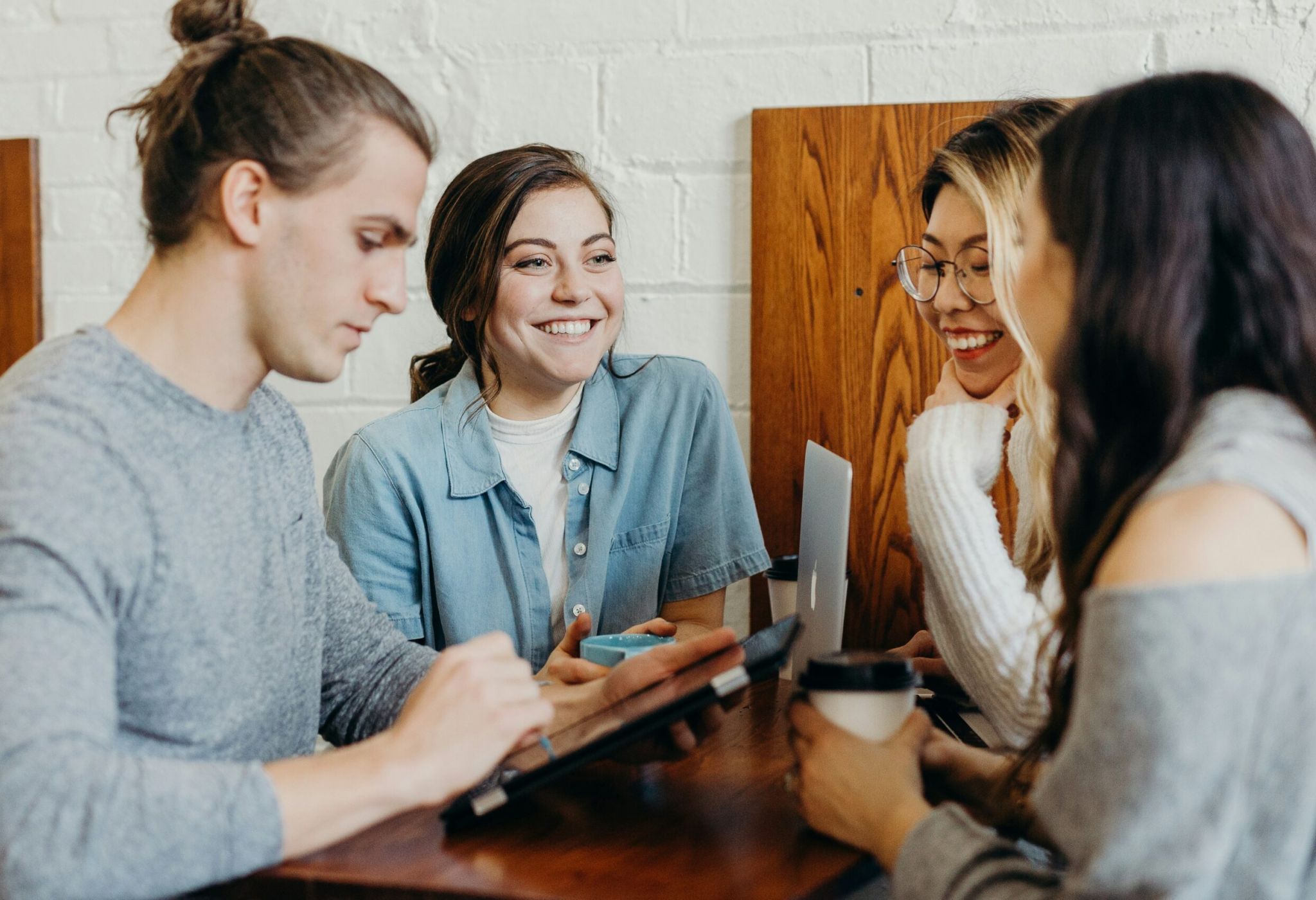 Four young adults sit at a table, chatting and smiling, with laptops and coffee cups in front of them.