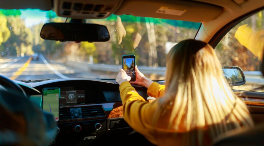 A woman in a car takes a photo with her phone while riding on a sunny road through a forest.