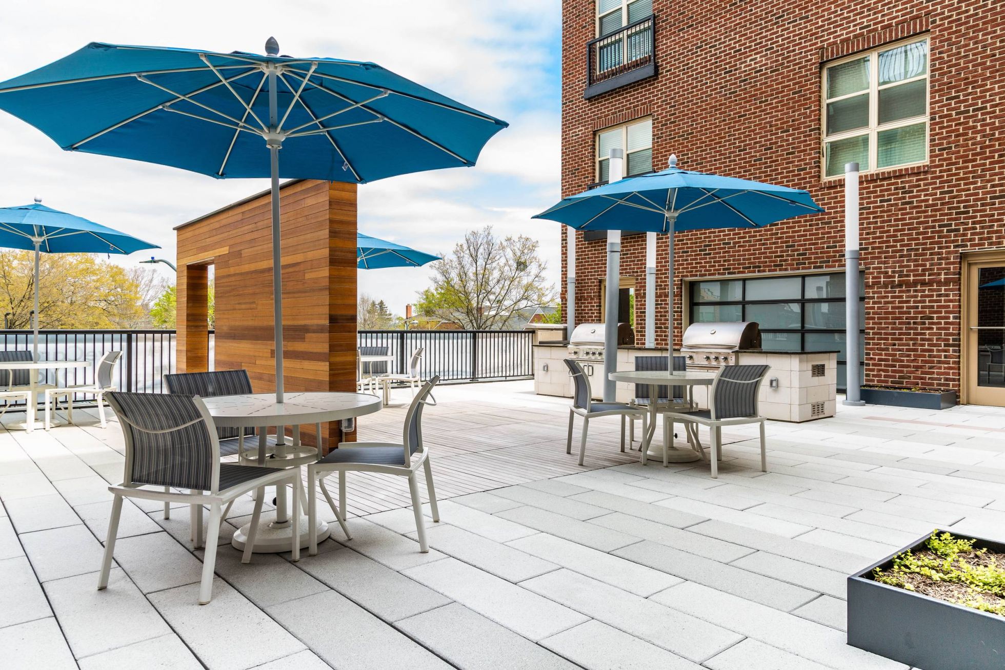 Outdoor patio with tables, chairs, blue umbrellas, and grills next to a brick building on a sunny day.