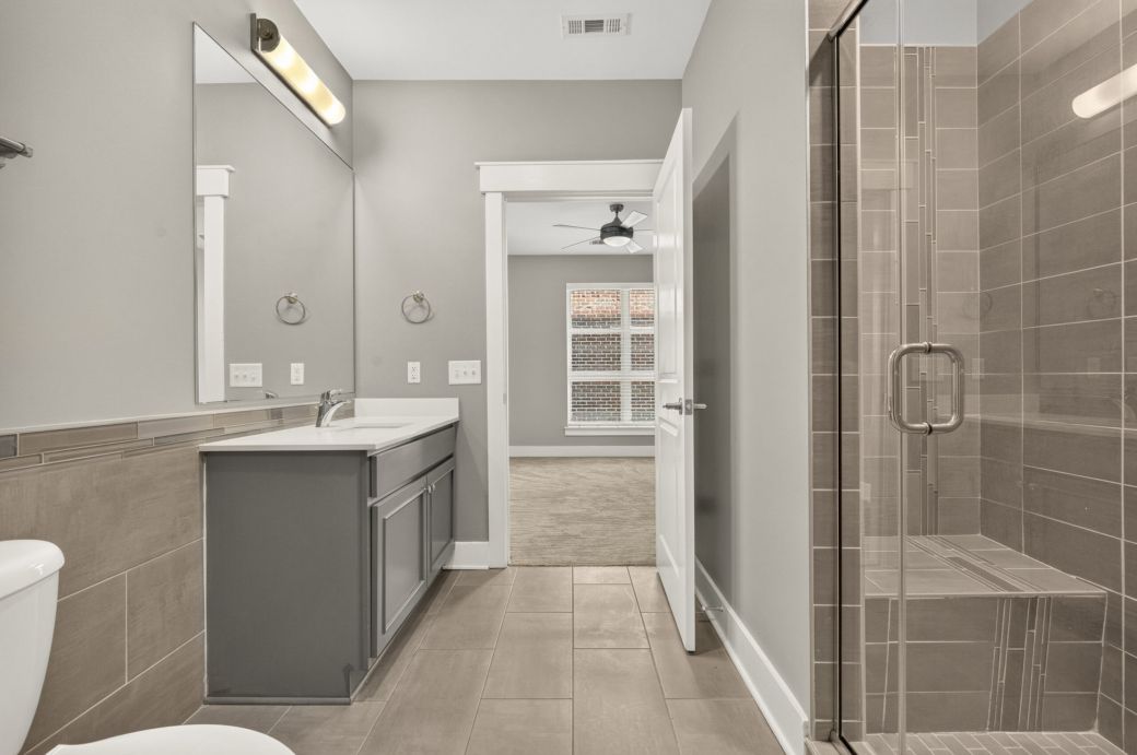 Modern bathroom with a glass shower, gray vanity, large mirror, and tile floor, leading to a bright bedroom with carpet.