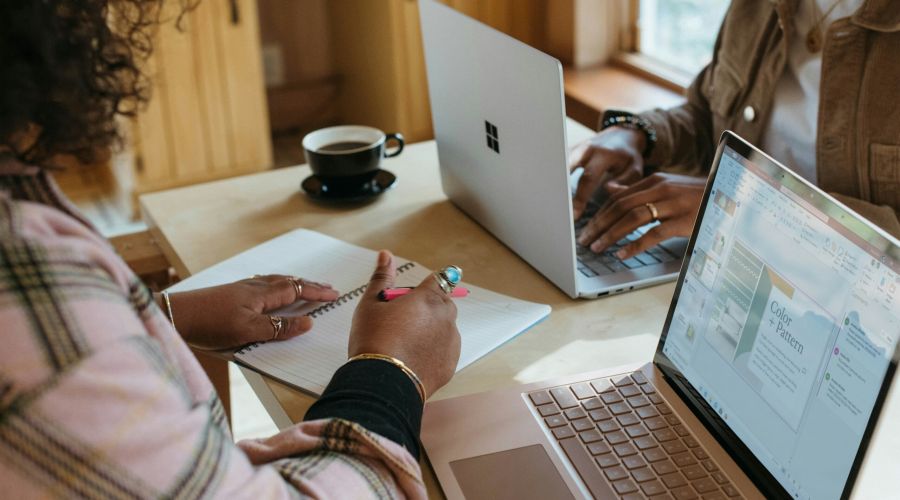 Two people working at a table with laptops and notebooks, one writing and the other typing.