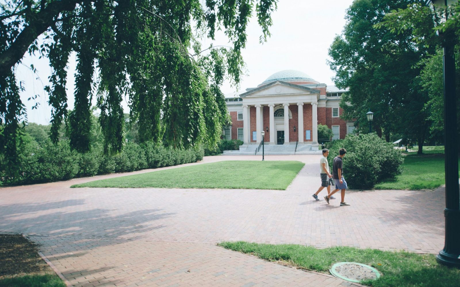 Two people walk on a brick path in front of a large, domed, brick building surrounded by trees.