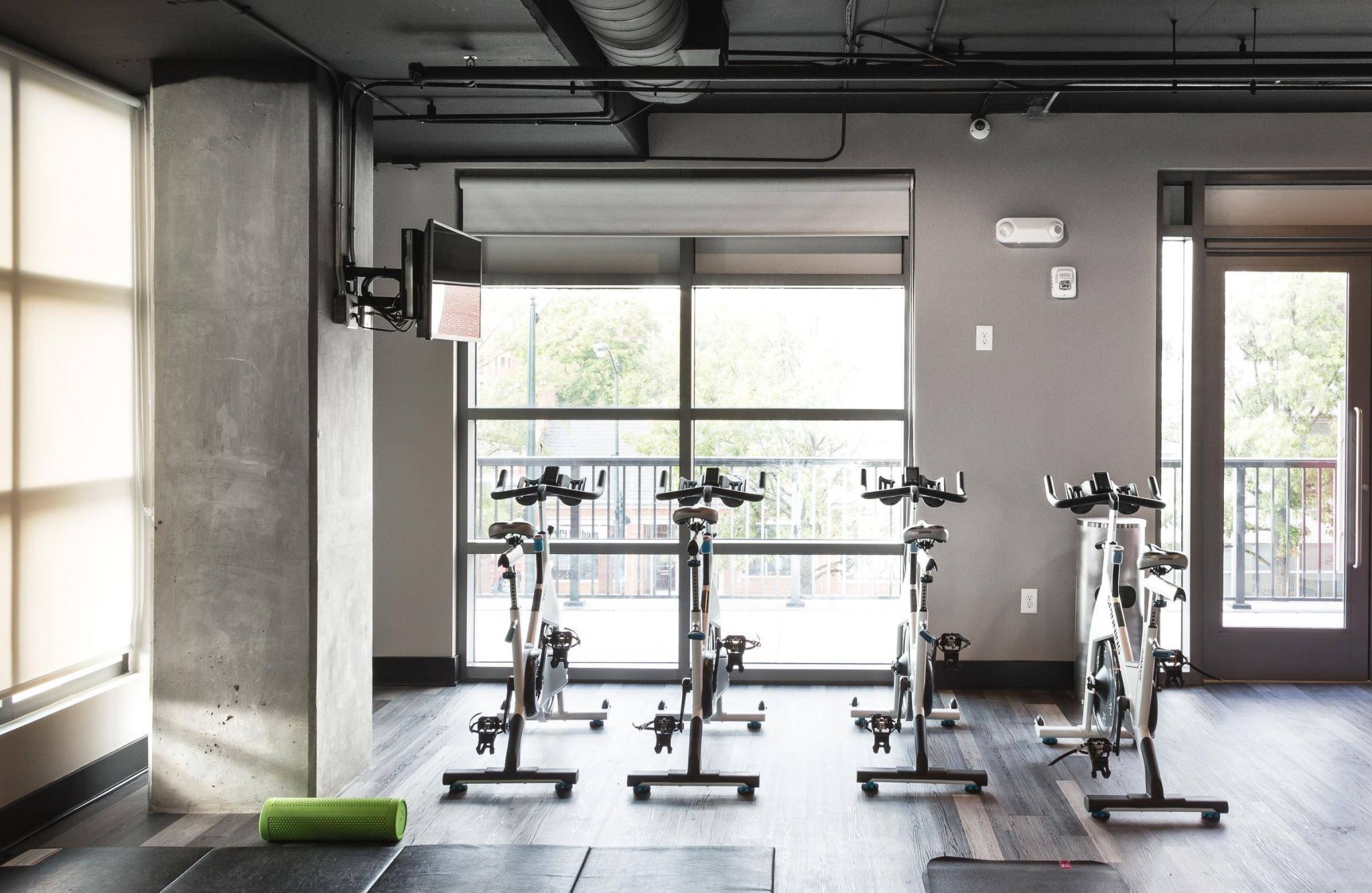 Three stationary bikes in a modern gym with large windows and a green foam roller on the floor.