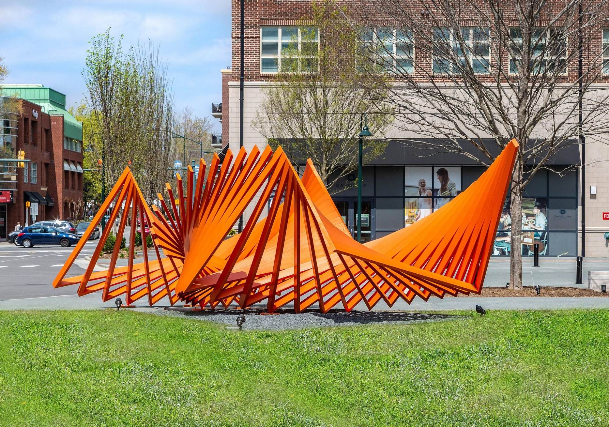 Bright orange metal sculpture with angular, overlapping beams on a grassy area in an urban setting.