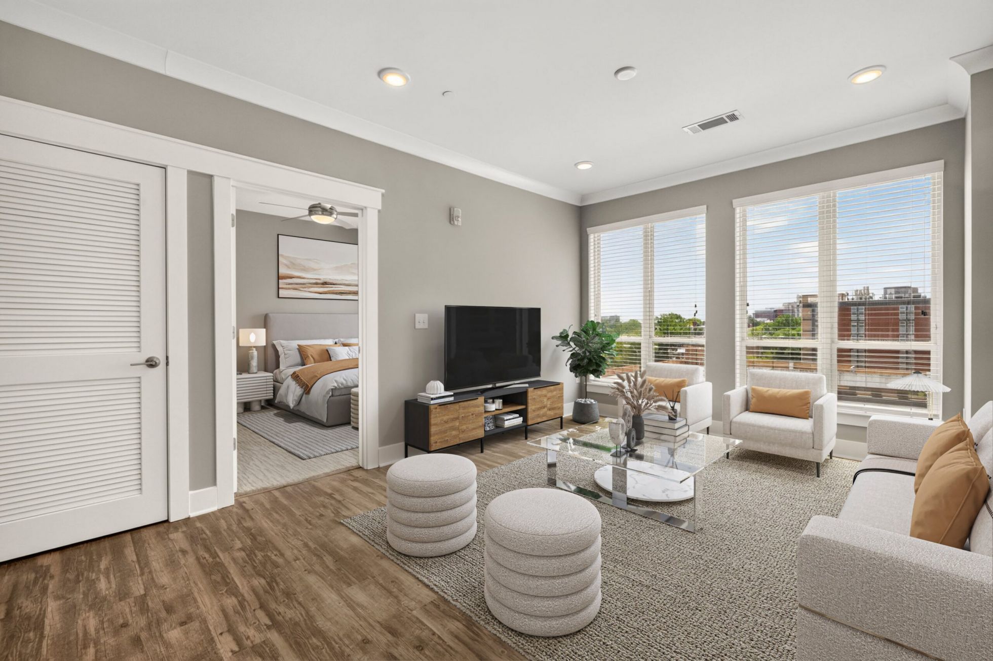 Modern living room with neutral decor, large windows, TV, two white chairs, and a view into a bedroom with a bed and artwork.